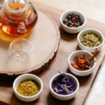 Glass teapot with herbal tea and assorted dry herbs in bowls on a wooden surface for aromatherapy.